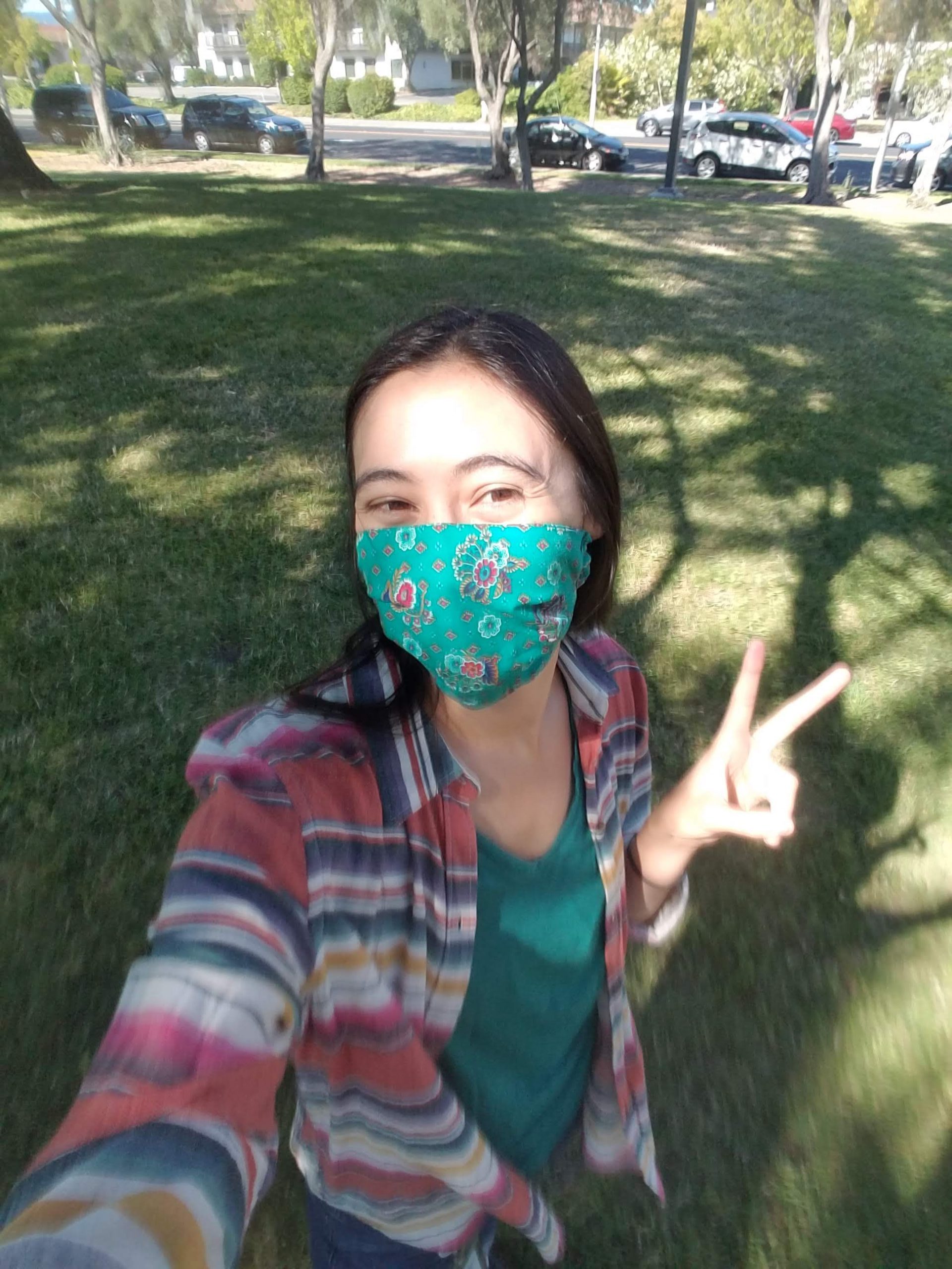 Woman in mask takes selfie in a yard while giving peace sign