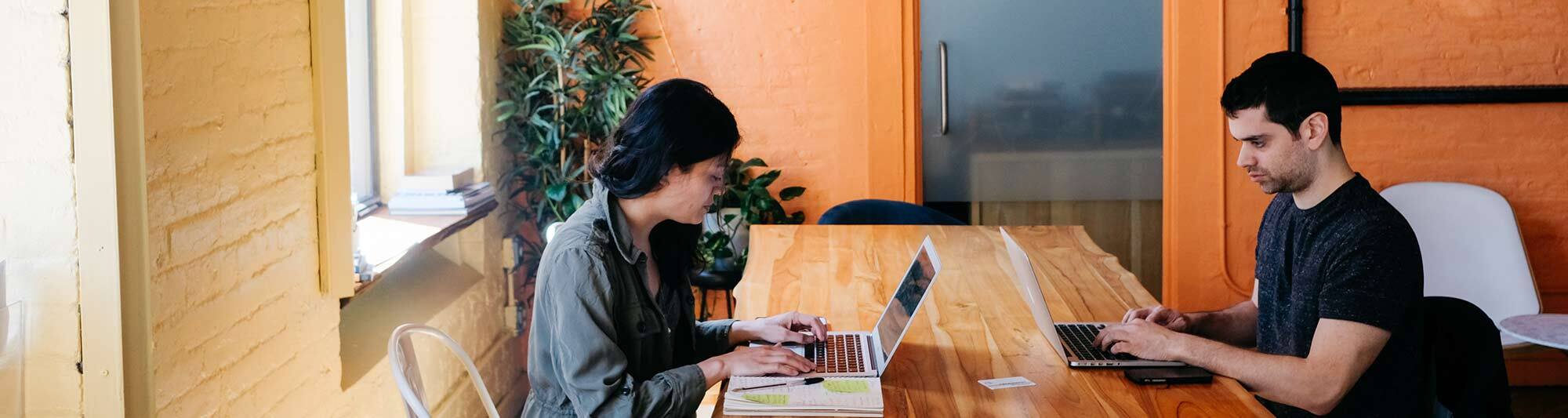 A woman and a man work on their computers while sitting across from each other at a long wooden table