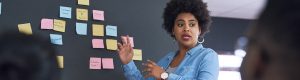 An African American woman stands in front of a board covered in multi-colored sticky nots, speaking to an audience