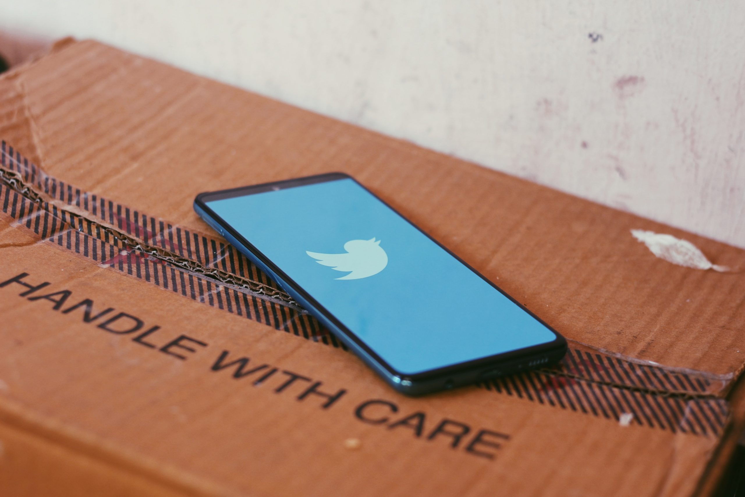 A smartphone displaying the Twitter logo against a light blue background sites atop a cardboard box labeled "handle with care."