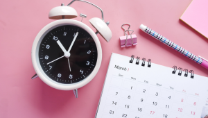 Looking down on soft pink background with calendar, binder clip, pencil, pink sticky note pad, and white alarm clock with a black face.