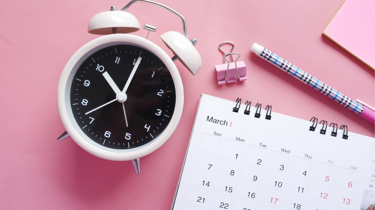 Looking down on soft pink background with calendar, binder clip, pencil, pink sticky note pad, and white alarm clock with a black face.