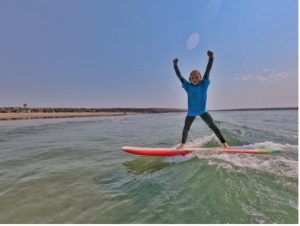 Child standing with hands above head on a surf board in the ocean.