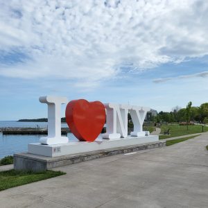 A statue reads "I heart NY" on a waterfront.