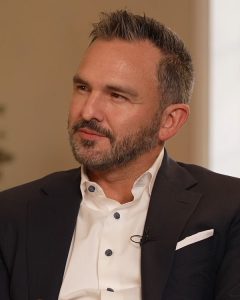 Head shot of a white man with brown, short hair and a mustache and beard. He is wearing a white shirt and dark blazer.