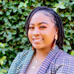 Headshot of black woman with shoulder length braids, wearing a blue plaid blazer with foliage in the background.