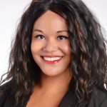 A headshot of a black woman, wearing a dark suit, standing against a light gray background