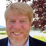 Headshot of white man with light red/blond hair and matching mustache/beard. Wearing a light dress shirt and dark blazer. He is standing outside near grass and a dark, leafy tree.