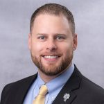 Headshot of white man against gray background. He has short, dark hair and a matching beard/mustache, and is wearing a light blue dress shirt, yellow tie, and dark blazer.