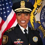 Headshot of black woman wearing dress police uniform, including hat, posing in front of an American flag and what looks like a police department flag.