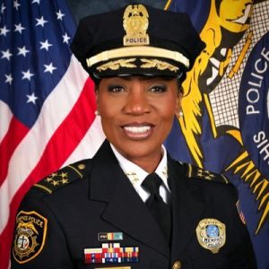 Headshot of black woman wearing dress police uniform, including hat, posing in front of an American flag and what looks like a police department flag.