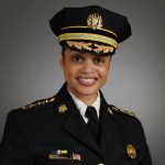 A headshot of a black woman wearing a police uniform, standing against a gray background