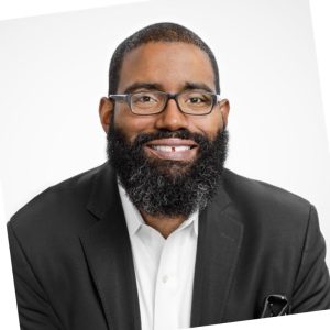 A headshot of a black man wearing glasses, a dark suit coat and a collared shirt, standing against a white background