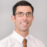 Headshot of white man with short dark hair and glasses, wearing a white dress short and orange-ish tie.