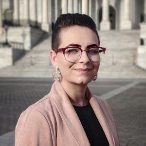 White person with short dark hair and glasses, a dark shirt and light pinkish blazer, and appears to be outside.