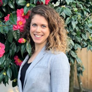 Portrait of white woman with shoulder length curly brown hair, wearing a dark shirt and light blue blazer, with flowers and greenery in the background.