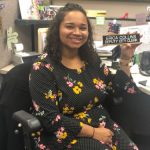 Brown skinned woman sitting at a desk, facing away from it and holding up a name plate that says Erica Collins, Deputy City Clerk.