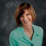 Headshot of white woman with a brown bob, wearing a mint green blazer and in front of a gray background.