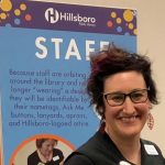 A headshot of a white woman with short hair, wearing glasses and a dark top, standing in front of a background with a placard that reads "Hillsboro Public Library Staff" above smaller text