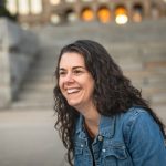 Headshot of while woman with long, dark brown, wavy hair. Looks like she is sitting on steps. She is wearing a denim jacket.