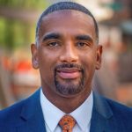 Headshot of black man with short black hair and goatee, wearing a light dress shirt, red tie, and blue blazer.