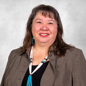 Headshot of woman with brown hair past her shoulders and bangs, wearing a dark short, brown blazer, and beaded necklace.