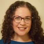 Headshot of white woman with shoulder length curly hair and glasses, wearing a dark top.