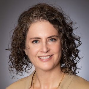 A headshot of a white woman wearing a tan suit and necklace, standing against a gray background