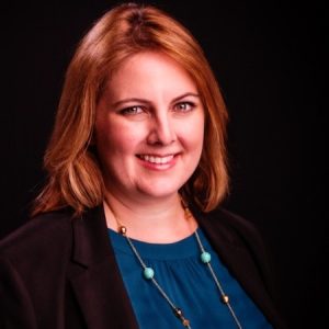 Headshot of white woman with shoulder length auburn hair, a blue top, black cardigan, against a black background.