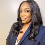 A headshot of a black woman wearing a dark blue suit and white shirt, standing against a beige background