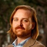 Headshot of white man with shoulder length auburn hair, beard and mustache, wearing a light colored dress shirt and brown blazer.