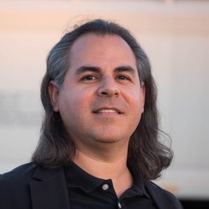 Headshot of white man with shoulder length dark grayish hair, and dark shirt, with a pastel colored background.