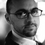 A black-and-white headshot of a white man, wearing glasses and a collared shirt