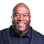 Headshot of black man with bald head and black dress shirt and blazer, against a white background.