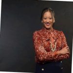 Asian woman standing with arms crossed, most hair pulled back, wearing dark reddish blouse.