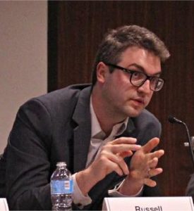 A photo of a white man, wearing glasses, seated at a table and gesturing with his hands toward a microphone