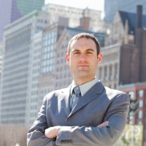 Portrait of white man with short dark hair, with arms crossed, wearing a light colored dress shirt, tie, and gray blazer, standing outside with city buildings behind him.