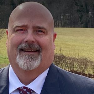 Man with bald head and gray goatee, wearing a light colored dress shirt, red tie, and dark blazer, standing outside.