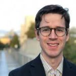 A headshot of a white man with dark hair, wearing glasses, a suit coat and collared shirt, standing against a background of a river and city