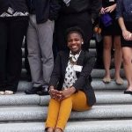 Black woman sitting on steps among other people standing. She's wearing mustard pants, a light blouse, and a dark blazer.