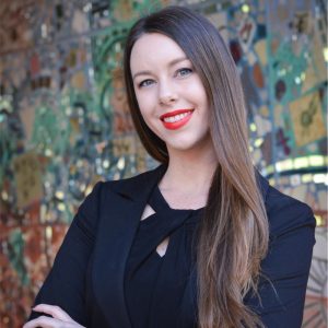 Portrait of a while woman with long brown hair, red lipstick, and a black top. She has her arms crossed and appears to be posing outside.