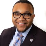 Headshot of black man with short dark hair and beard, glasses, wearing a light colored dress shirt, colorful tie, and dark blazer.