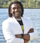 Black man standing with arms crossed in front of a body of water. He has shoulder-length dark hair and is wearing a white blazer.