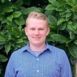 Headshot of white man with short, blond hair. Wearing a blue button-up shirt and standing in front of dark green leafy vegetation.