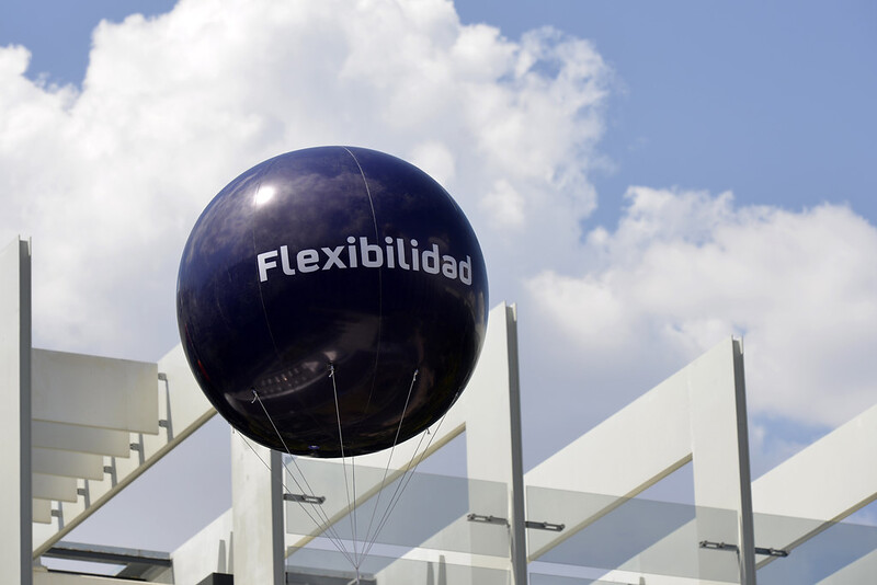 A navy sphere that says Flexibilidad in white is floating with some open, white architecture, against a blue sky with white fluffy clouds.