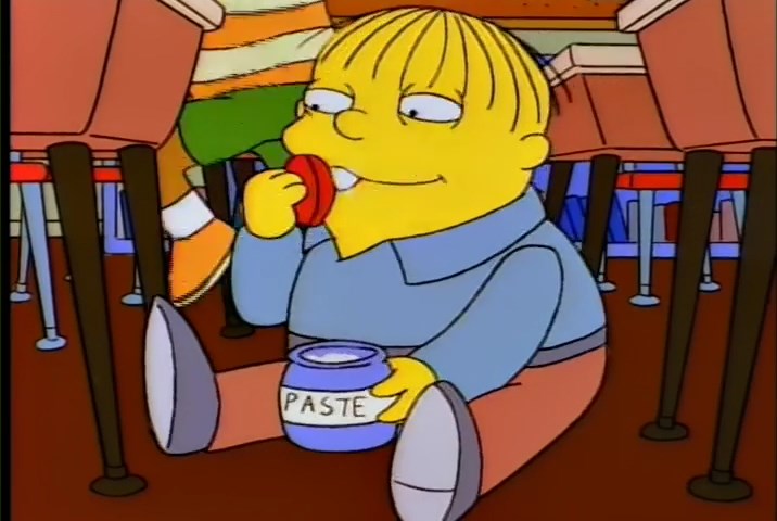 The character Ralph from The Simpson's, sitting on a classroom floor, eating from a paste jar.