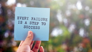 Blurred outside background. Hand in foreground holds a blue sticky note with typed words "every failure is a step to success."