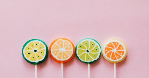 Light pink background with four lollipops sticking up from the bottom, in orange, green, blue, yellow colors.