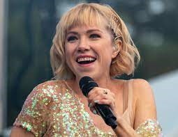 Image of singer Carly Rae Jepson with blond hair, gold sequin top, and holding a microphone.