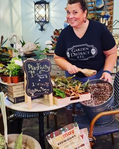 A woman wearing a black T-shirt holds a metal garden planter inside a gardening shop filled with plants and gardening supplies.